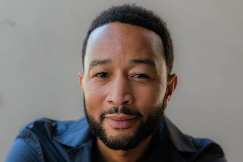 A portrait of John Legend sitting in a chair with his arms crossed leaning on the back of the chair